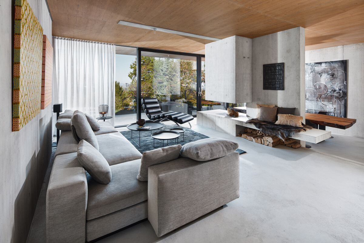 Beton, Holz und Glas bilden die Basis des modernen Neubaus. Mittelpunkt ist das offen gestaltete Erdgeschoss mit Wohn-, Koch- und Essbereich. Eine Besonderheit ist bereits von außen gut ersichtlich – das verbindende Element Eichenholz. Im Erdgeschoss ziert dieses die Decke und im Obergeschoss den Boden. Der Einsatz von massivem Holz, sowie hochwertigen Textilien und bunten Kunstwerken verleihen dem Beton Leben und Natürlichkeit. Die Einrichtung ist in klaren Grau- und Schwarztönen gehalten. Im Außenbereich werden diese Farbtöne durch ein kräftiges Pink als Blickfang ergänzt.